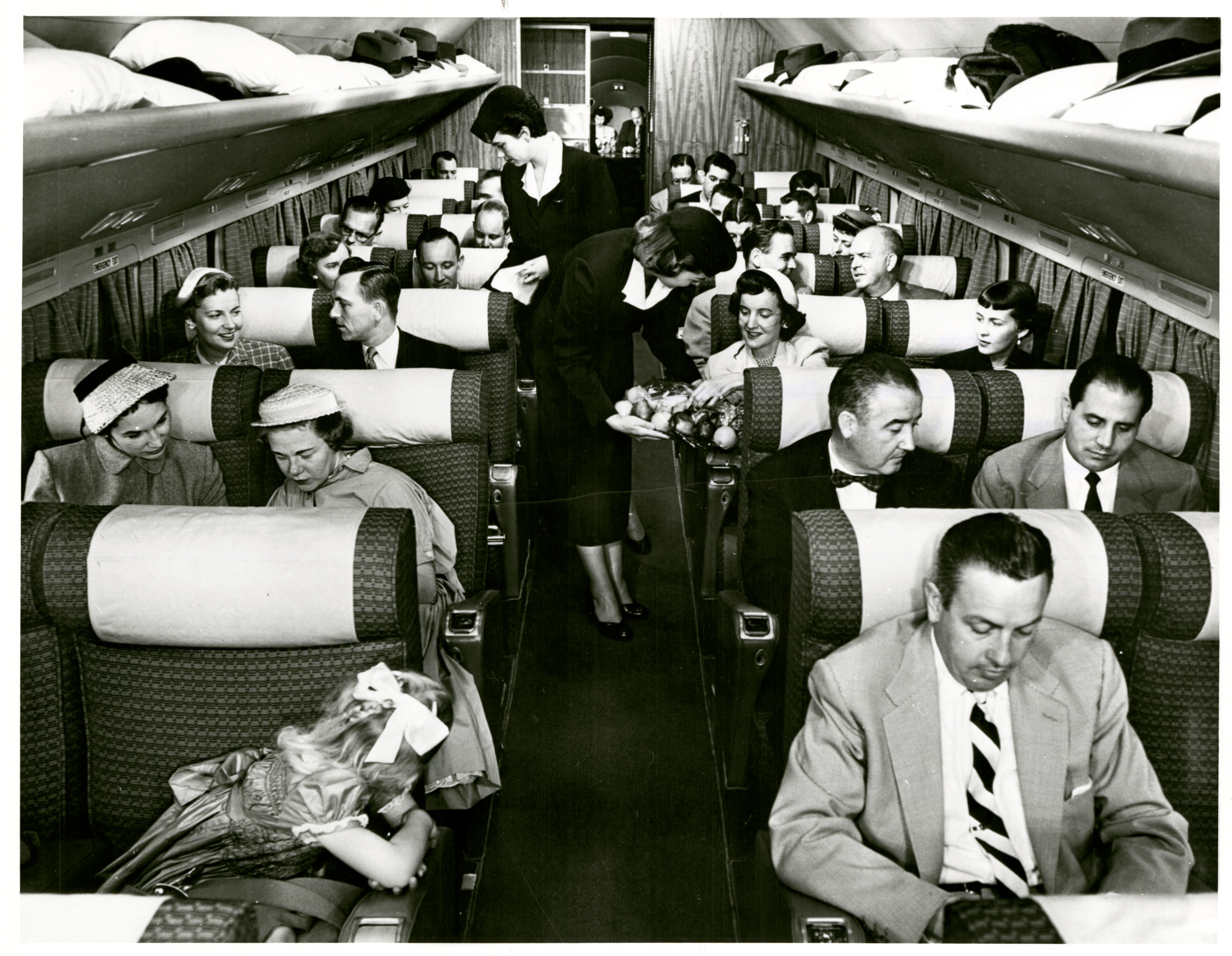 Photograph of a full airplane cabinet, with stewardesses walking the aisles serving the passengers.
